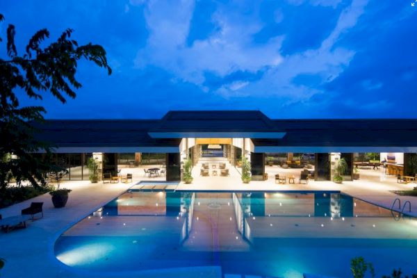 A luxurious, modern building with a large, illuminated swimming pool in front, captured at dusk with a deep blue sky.