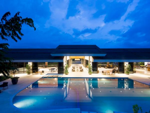 A luxurious, modern building with a large, illuminated swimming pool in front, captured at dusk with a deep blue sky.