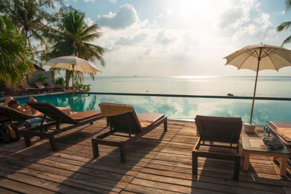 Poolside wooden deck with lounge chairs, umbrellas, and palm trees overlooking a calm ocean with a bright sky and scattered clouds ending the sentence.