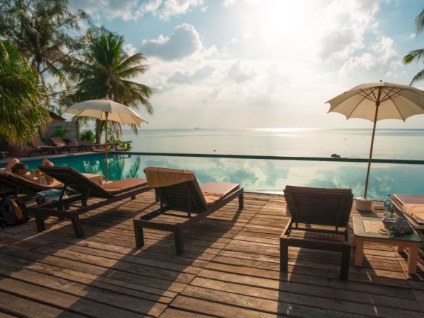Poolside wooden deck with lounge chairs, umbrellas, and palm trees overlooking a calm ocean with a bright sky and scattered clouds ending the sentence.