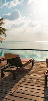 A serene poolside scene with lounge chairs, umbrellas, palm trees, and a tranquil view of the ocean under a bright sky, perfect for relaxation.