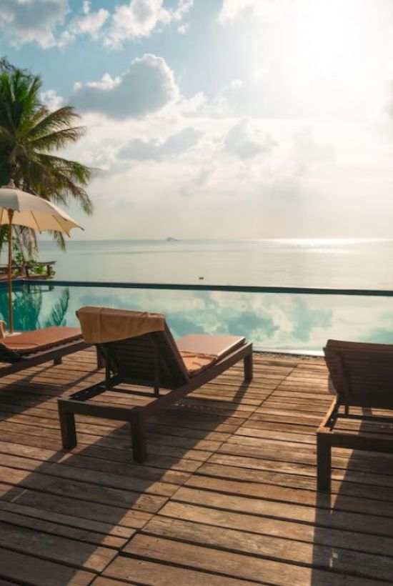 An inviting poolside setting with lounge chairs and umbrellas facing a serene, sunlit ocean view, flanked by palm trees and a wooden deck.