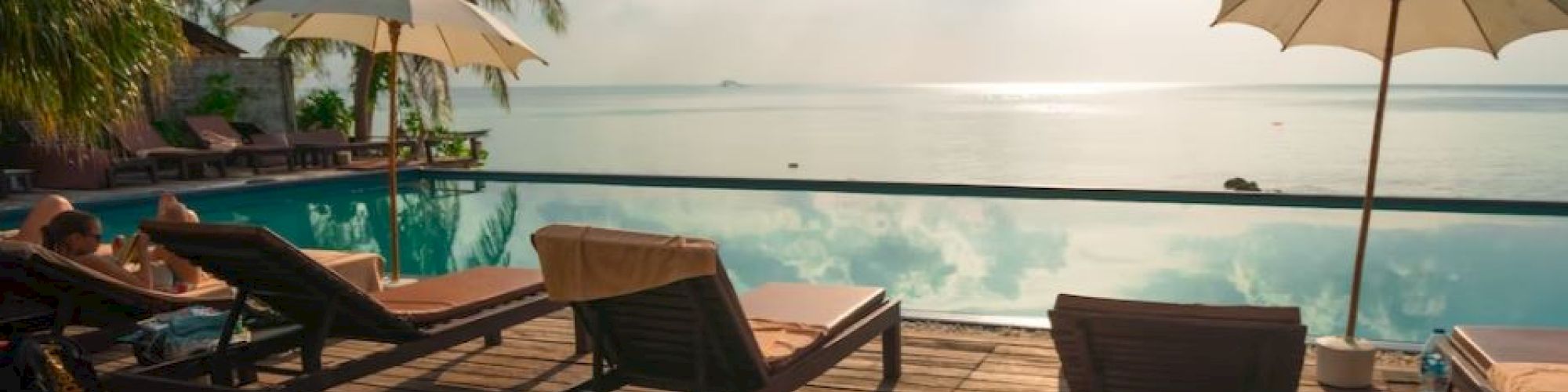 A serene poolside scene with lounge chairs and umbrellas overlooking a calm ocean, palm trees, and a bright sun reflecting off the water.