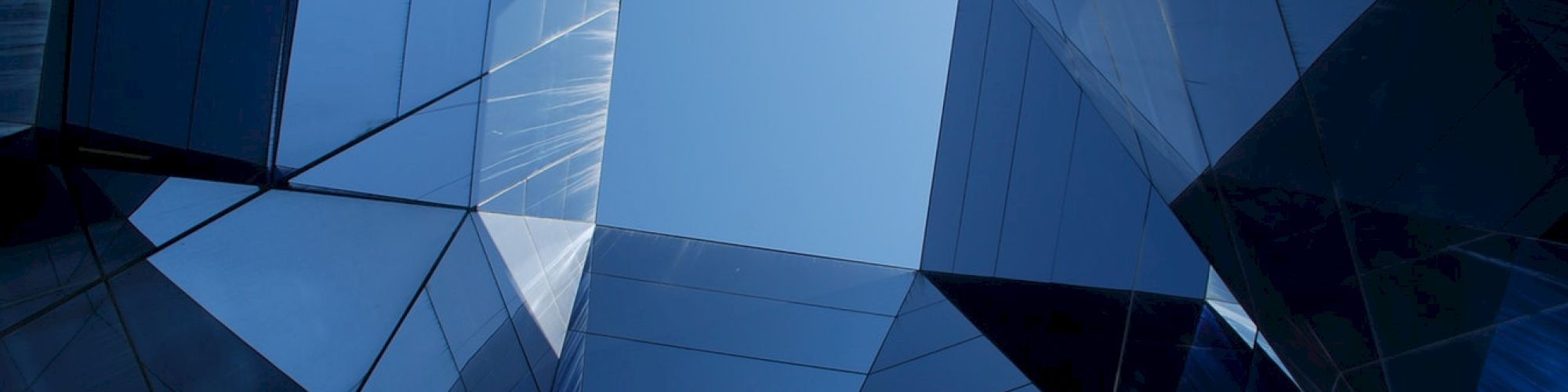 The image shows an abstract perspective looking up between angular glass buildings, forming a geometric pattern with a patch of blue sky at the center.