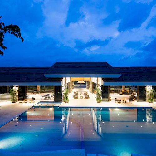 A luxurious modern building with a large illuminated swimming pool at night, featuring open-air seating and surrounded by a serene environment.