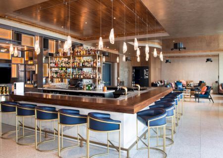 A modern bar with a variety of drinks, blue barstools, stylish lighting, and an inviting lounge area in the background.