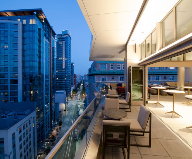 This image features an outdoor balcony with seating and tables, overlooking a city street with modern high-rise buildings lit up in the evening.