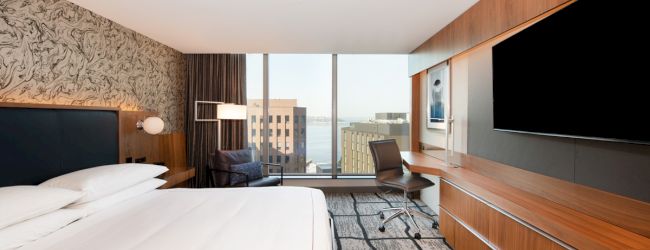 A modern hotel room featuring a large bed, wall-mounted TV, desk, chair, and a view of city buildings outside the window.