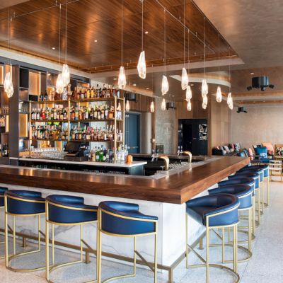 A modern bar with a fully stocked back shelf, high stools around a wooden counter, hanging lights, and a comfortable lounge area in the background.