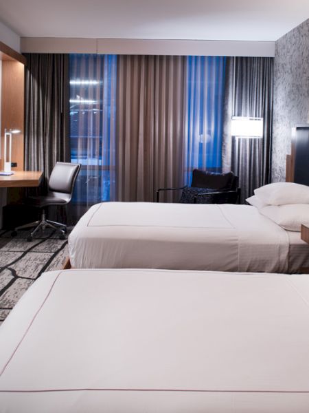 A modern hotel room with two beds, a desk, chair, lamp, flatscreen TV, and contemporary decor, featuring a patterned wall and large window draped with curtains.