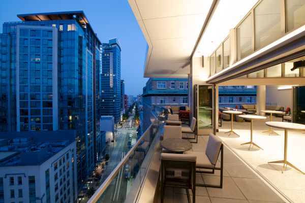 This image shows a modern outdoor balcony with seating and tables overlooking a cityscape with tall buildings at dusk.