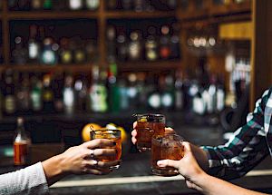 Three people raising their glasses in a toast in a dimly lit bar with shelves of bottles in the background.