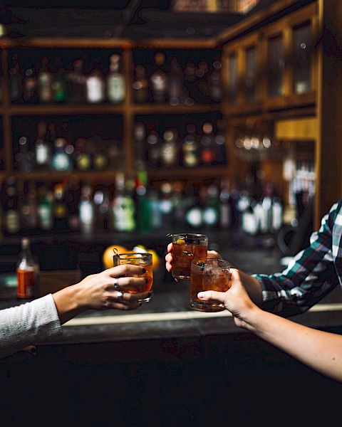 Three people raising their glasses in a toast in a dimly lit bar with shelves of bottles in the background.