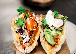 The image shows two tacos filled with various ingredients like meat, vegetables, and sauces, garnished with cilantro and placed on a white plate.