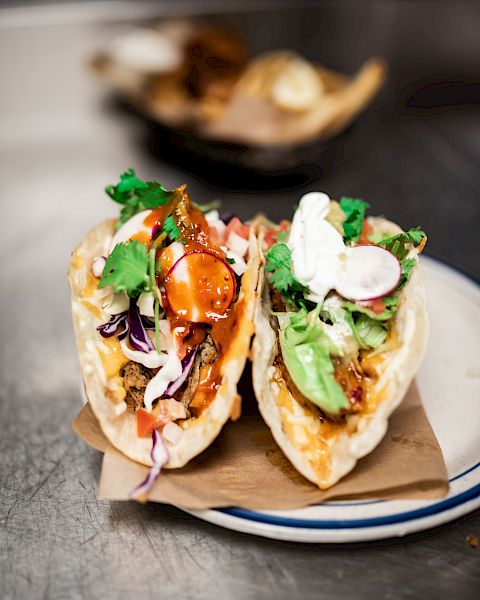 The image shows two tacos filled with various ingredients like meat, vegetables, and sauces, garnished with cilantro and placed on a white plate.