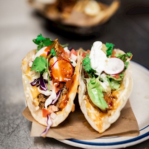 The image shows two tacos filled with various ingredients like meat, vegetables, and sauces, garnished with cilantro and placed on a white plate.