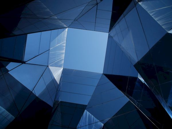 The image shows a geometrical arrangement of reflective glass structures, forming a skylight that opens up to a blue sky.
