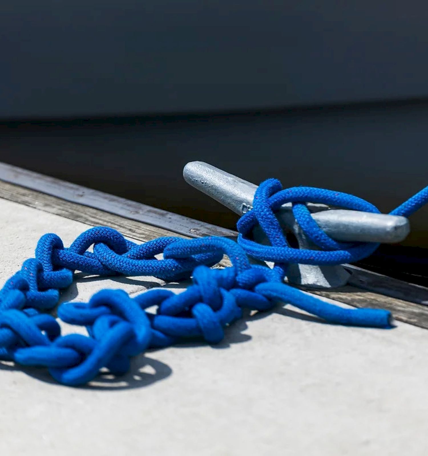 A coiled blue rope is secured to a cleat on a waterfront dock, with part of a boat visible in the background.