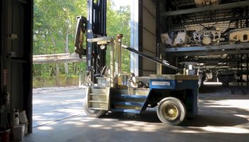 A forklift with extended forks is inside a warehouse, near a large open door, with some trees visible outside in the background.