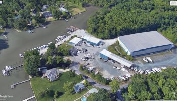 This image shows a marina with numerous boats docked, storage facilities, and a large building surrounded by a wooded area and nearby houses.