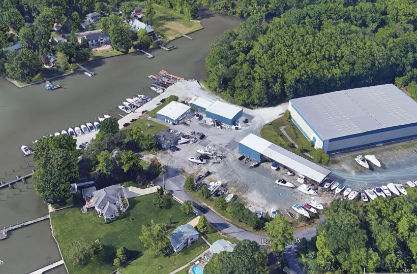 This image shows a marina with numerous boats docked, storage facilities, and a large building surrounded by a wooded area and nearby houses.