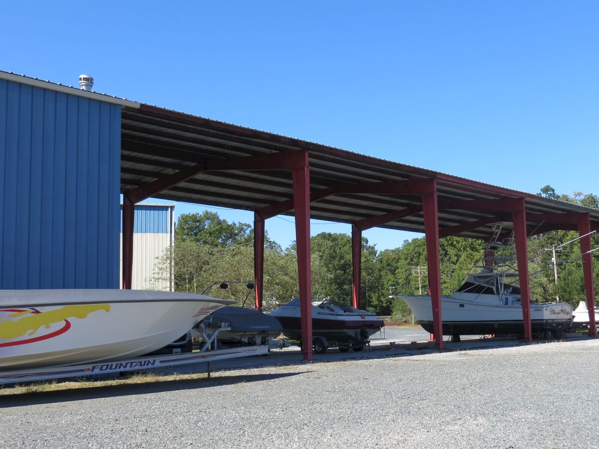 The image shows a covered boat storage area with several boats under a red metal roof, adjacent to a blue building, with a background of trees.