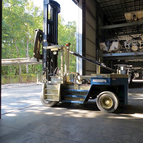 An industrial forklift is parked inside a warehouse near an open door, with sunlight and greenery visible outside.