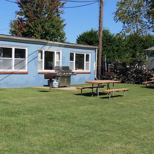 A grassy yard with picnic tables, a blue building with windows, a BBQ grill, and trees in the background under a clear blue sky.