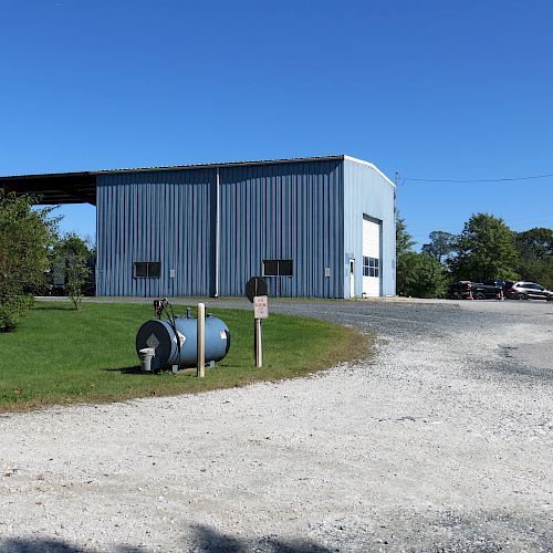 The image shows a blue industrial building surrounded by gravel with a fuel tank in the foreground, trees, and a clear blue sky, creating a rural setting.