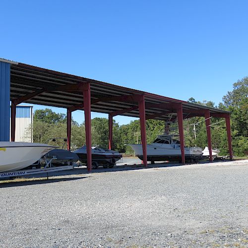 Several boats are stored under a red metal shed, next to a blue building, on a gravel surface, with trees in the background under a clear blue sky.