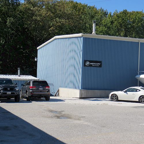 The image shows a blue metal building with a sign, three parked vehicles, and a boat on a trailer. The area is bright and sunny.