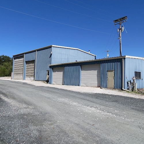 The image shows a few blue warehouse buildings with large garage doors, situated along a gravel road, under a clear blue sky, next to some greenery.