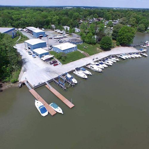 An aerial view of a marina with multiple boats docked along the shore, surrounded by trees, buildings, and parking. The water looks calm and peaceful.