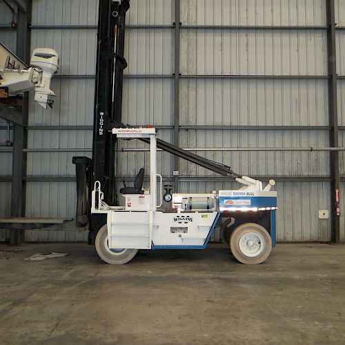 The image shows a white forklift inside a warehouse with large metal walls and a concrete floor. There is also a boat engine on the upper left side.