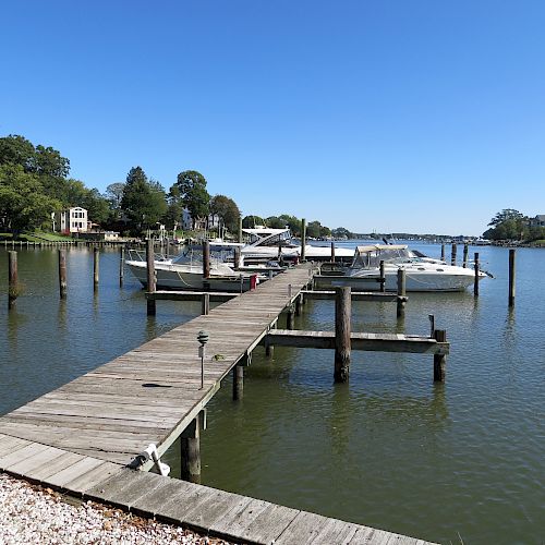 A wooden dock extends over calm waters, with several boats moored alongside, surrounded by a peaceful landscape and clear blue sky.