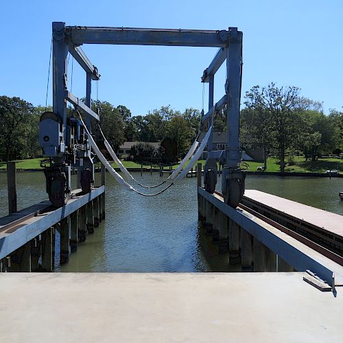 The image shows a boat lift at a waterfront, with trees and houses in the background across the water.