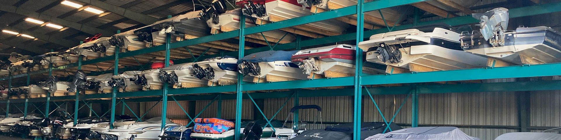 Stacks of boats on metal racks inside a warehouse, covered in covers and some on trailers awaiting storage or transport.