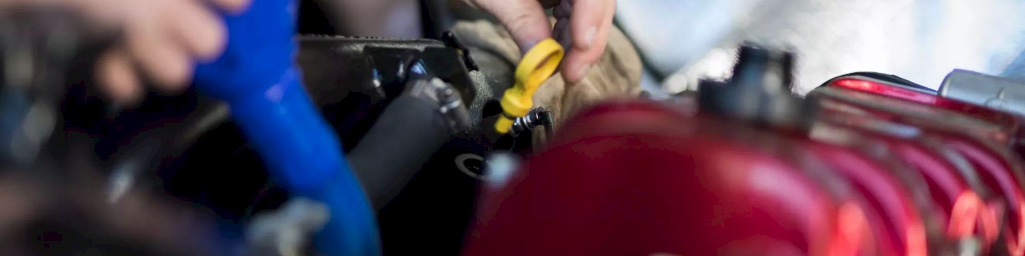 Someone pouring fluid into a car engine while another hand holds a yellow dipstick; red engine block in focus.