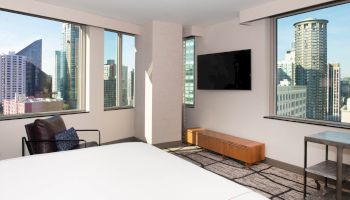 A modern hotel room with large windows offering city views, a flat-screen TV, a bed, an armchair, and a small table.