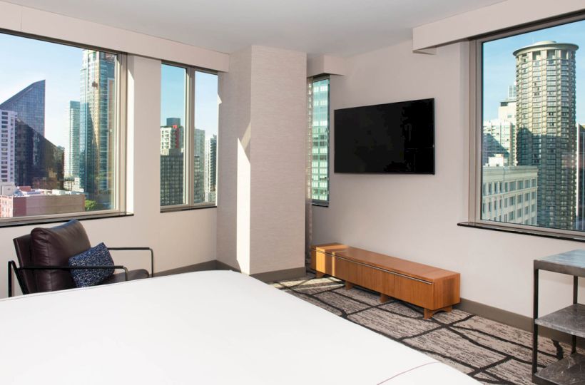 A modern hotel room with large windows offering city views, a flat-screen TV, a bed, an armchair, and a small table.