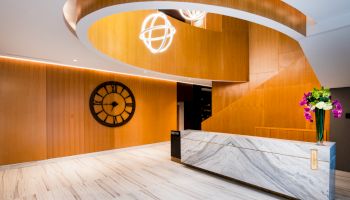 A modern lobby with wooden walls, a large wall clock, a marble front desk, decorative lighting, and a vase with flowers on the desk.