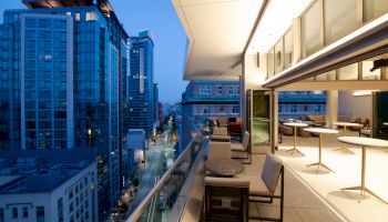 A modern balcony view overlooking a cityscape with tall buildings at dusk, featuring outdoor seating and tables on the balcony.