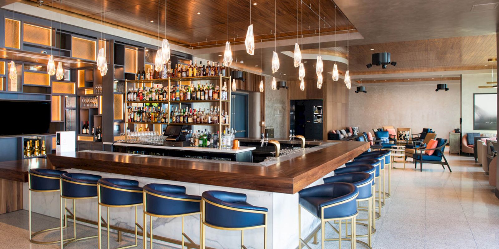 The image shows a modern bar with a wooden countertop, blue cushioned stools, hanging lights, a well-stocked liquor shelf, and a spacious seating area.