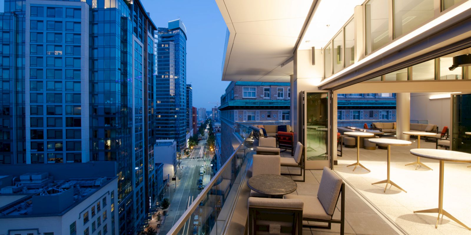 A modern balcony at night with tables and chairs overlooks an urban street filled with high-rise buildings and city lights.