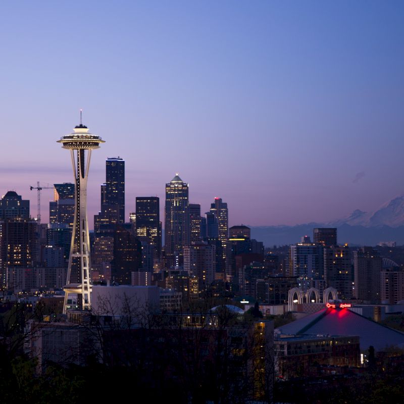 The image shows the Seattle skyline at dusk with the iconic Space Needle and Mt. Rainier visible in the background, bathed in twilight.