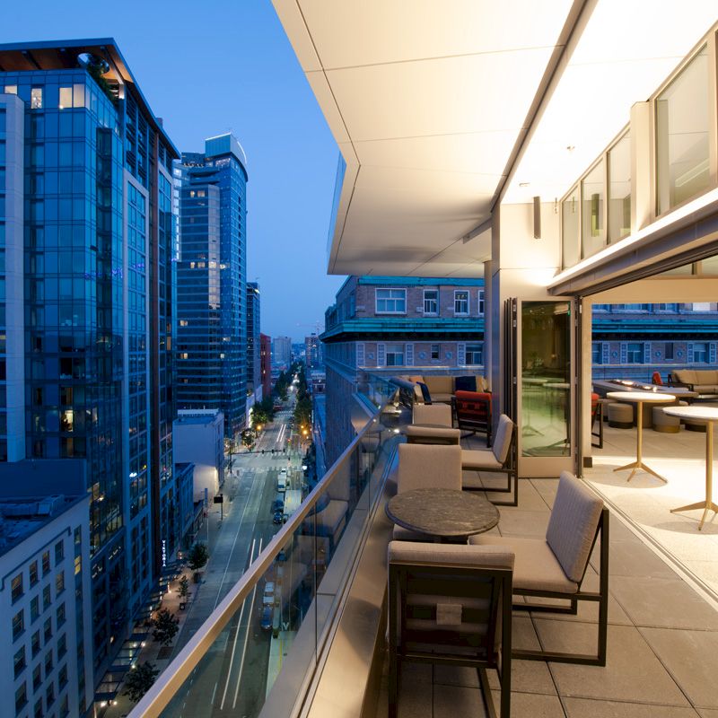 A modern balcony with tables and chairs overlooks a city street lined with tall buildings at dusk, with lights illuminating the scene.