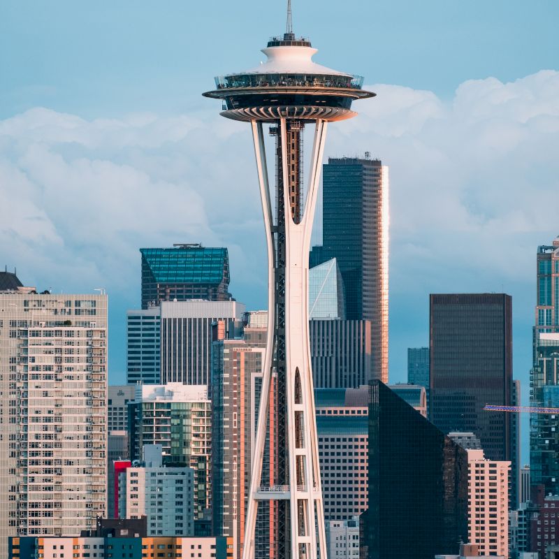 The image shows the Space Needle in Seattle, surrounded by various tall buildings and skyscrapers, with a backdrop of cloudy sky and an urban cityscape surrounding.