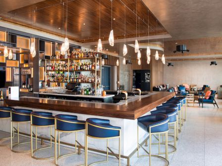 A modern bar with stylish seating, a well-stocked bar area with various bottles, hanging lights above the counter, and seating areas in the background.