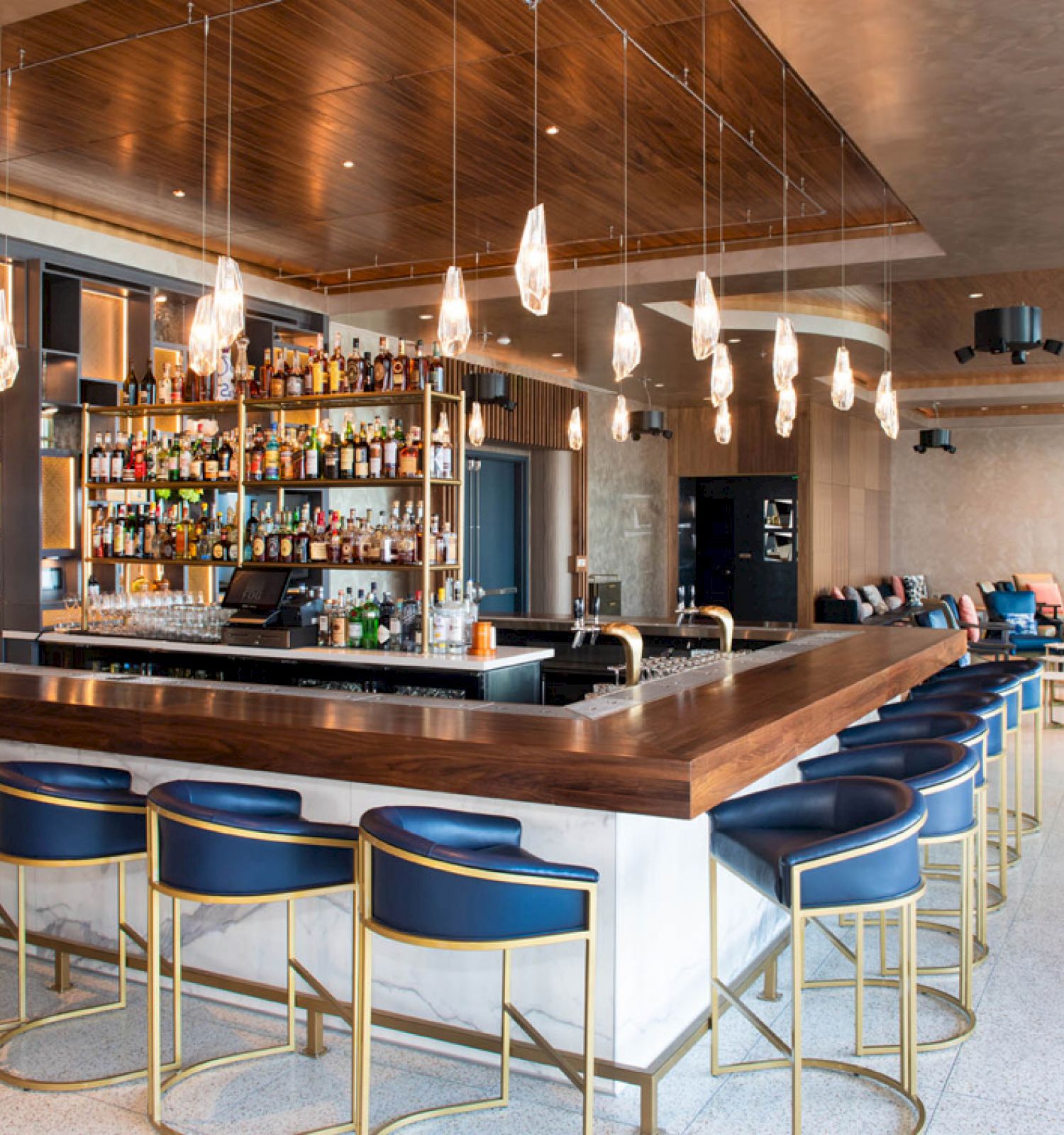 A modern bar with blue bar stools, hanging lights, and shelves stocked with bottles, surrounded by a spacious seating area and a neutral color palette.