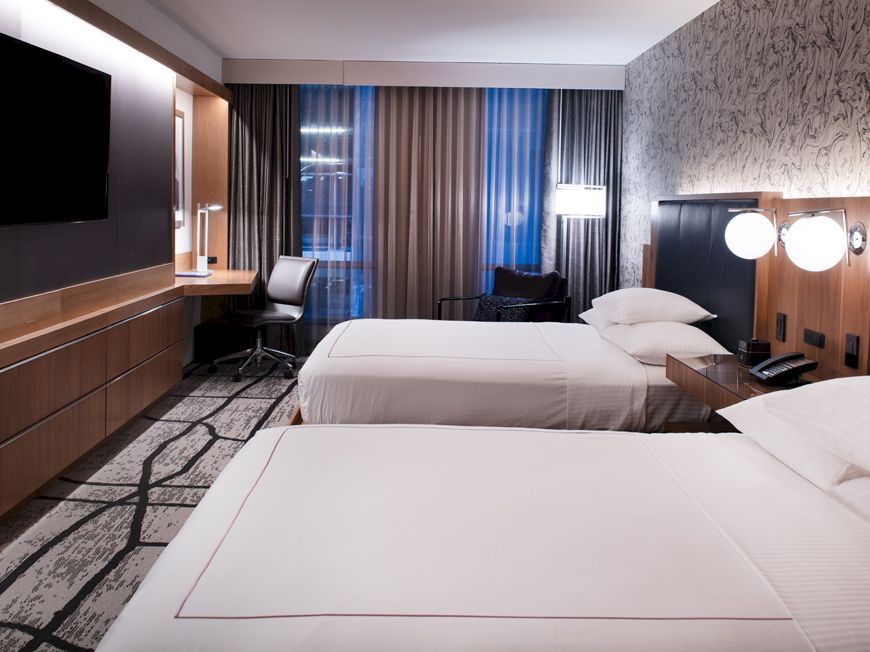 A modern hotel room with two beds, a wall-mounted TV, a work desk, and stylish decor, including patterned wallpaper and a carpet.